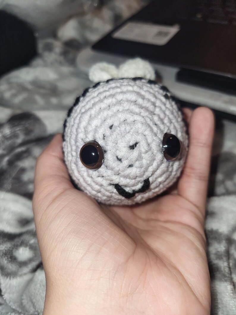 May include: A handmade crochet bee with a grey and white body, black stripes, and large brown eyes. The bee has a smiling face and small white wings. The toy is held in a hand.