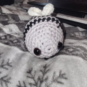 May include: A crocheted bee toy, predominantly white with black stripes. It features large, dark brown eyes and small white antennae. The toy is resting on a soft, grey blanket with a snowflake pattern.