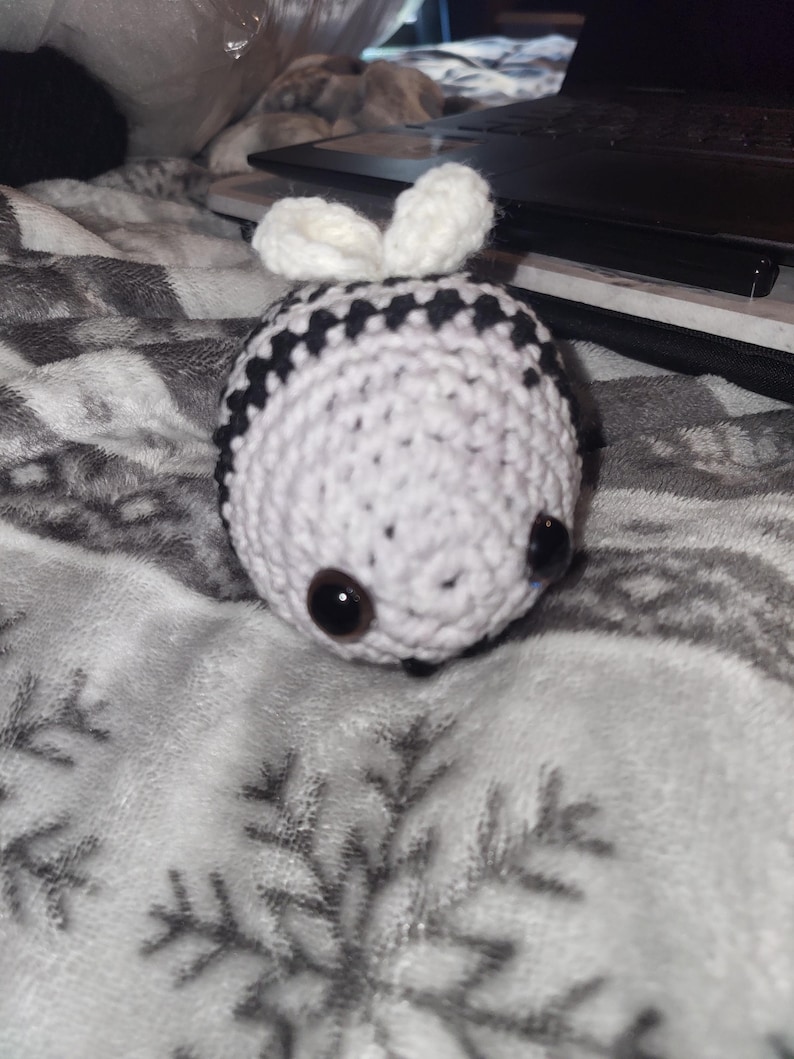 May include: A crocheted bee toy, predominantly white with black stripes. It features large, dark brown eyes and small white antennae. The toy is resting on a soft, grey blanket with a snowflake pattern.