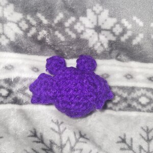May include: A handmade, purple crochet bat with a textured appearance. The bat has two small ears and outstretched wings. The bat is resting on a soft, gray and white snowflake patterned blanket.