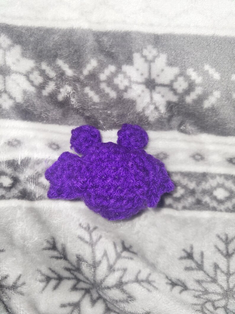 May include: A handmade, purple crochet bat with a textured appearance. The bat has two small ears and outstretched wings. The bat is resting on a soft, gray and white snowflake patterned blanket.