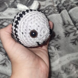 May include: A crocheted bee toy, predominantly white with black stripes, featuring large brown eyes and small white wings. The bee is held in a hand, set against a grey patterned backdrop.