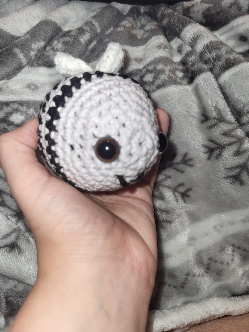 May include: A crocheted bee toy, predominantly white with black stripes, featuring large brown eyes and small white wings. The bee is held in a hand, set against a grey patterned backdrop.