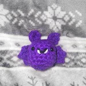 May include: A handmade purple crochet bat with small black eyes and white fangs. The bat has large ears and outstretched wings. The bat is sitting on a gray and white snowflake patterned blanket.
