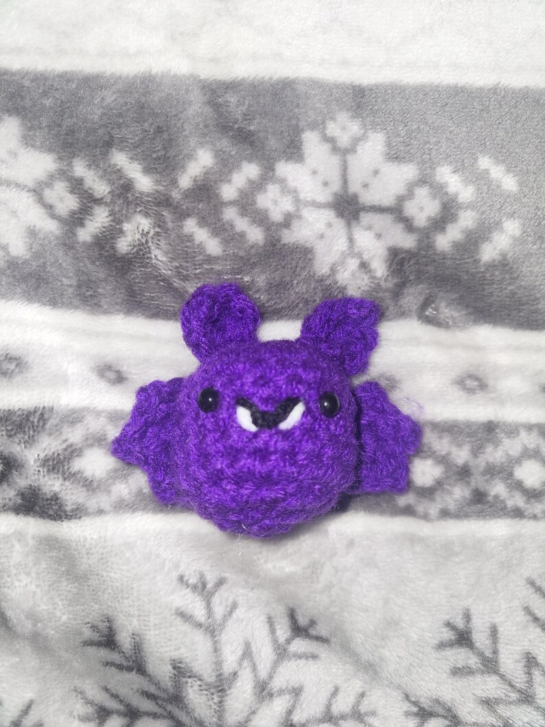 May include: A handmade purple crochet bat with small black eyes and white fangs. The bat has large ears and outstretched wings. The bat is sitting on a gray and white snowflake patterned blanket.