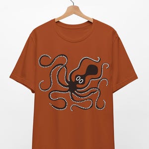 May include: Rust-colored t-shirt featuring a black octopus design. The octopus has white dotted accents along its tentacles and a simple, cartoon-like face. The shirt is hanging on a wooden hanger.