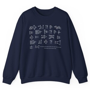 May include: A navy blue crewneck sweatshirt featuring a white graphic print of cuneiform script. Below the symbols, the text reads: If one destroys the sight of another, their sight shall be destroyed. Hammurabi's Law 196: Eye for an eye.