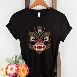 May include: Black t-shirt featuring a detailed graphic of a stylized face with three eyes, red and gold accents, and a wide open mouth. The design is centered on the front of the shirt. The shirt is hanging on a wooden hanger.