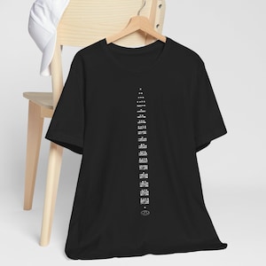 May include: A black short-sleeved t-shirt featuring a vertical column of white text and small circular icons arranged in a pyramid-like sequence down the centre of the chest, accompanied by a plain white baseball cap hanging on a wooden chair.