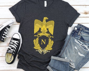 May include: A dark charcoal gray short-sleeve t-shirt featuring a yellow graphic of an eagle perched above a letter N surrounded by a laurel wreath. The shirt is displayed alongside a pair of black low-top sneakers and distressed blue jeans.
