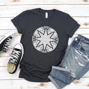 May include: A dark charcoal grey short-sleeved t-shirt featuring a white circular graphic with a distressed texture and a central eight-pointed star design. The shirt is displayed alongside a pair of black low-top trainers and blue denim jeans.