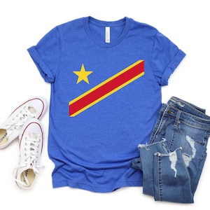 May include: A blue short-sleeve t-shirt featuring a yellow star and a diagonal red stripe bordered in yellow, paired with white canvas sneakers and blue denim jeans with distressed details on the legs and pockets.