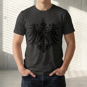 Puede incluir: Camiseta gris oscuro con un diseño de águila heráldica negra. El gráfico incluye una corona, alas y detalles ornamentados. La camiseta es de cuello redondo y parece estar hecha de un material suave.