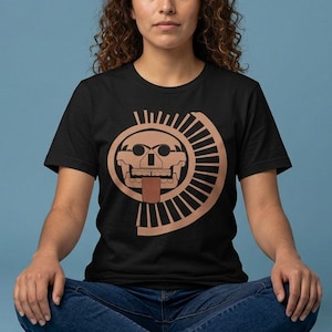 Op de afbeelding: Zwart T-shirt met een bruin schedelontwerp en een zonachtige spiraal. De grafische tee heeft een ronde hals en wordt gedragen door een persoon die met gekruiste benen zit. De achtergrond is effen blauw.