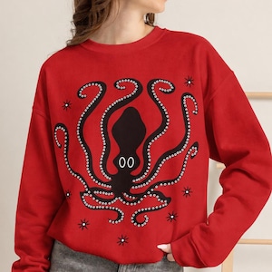 May include: A bright red long-sleeve sweatshirt featuring a large black octopus graphic centered on the front. The octopus has white circular eyes and is surrounded by small black star-shaped accents. The sweatshirt has a crew neckline.
