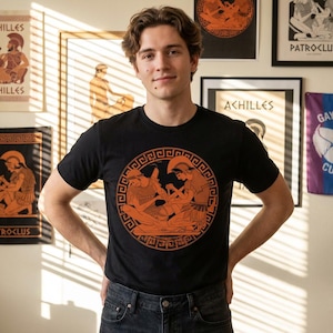 May include: Black t-shirt featuring an orange illustration of Greek figures within a circular design. The artwork depicts two figures in helmets and armor. The shirt is worn by a person in front of a wall with framed art and posters.