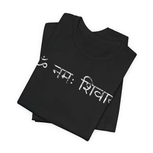 May include: A black t-shirt featuring white distressed text printed diagonally across the front. The text is written in Devanagari script and reads Om Namah Shivaya. The shirt is folded, showing a crew neckline and short sleeves.