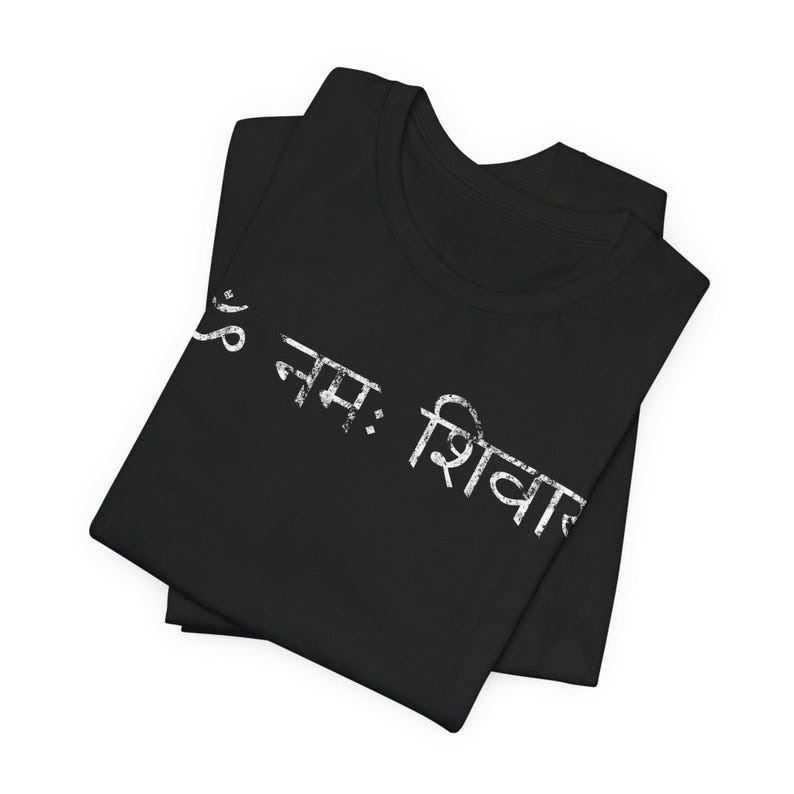 May include: A black t-shirt featuring white distressed text printed diagonally across the front. The text is written in Devanagari script and reads Om Namah Shivaya. The shirt is folded, showing a crew neckline and short sleeves.