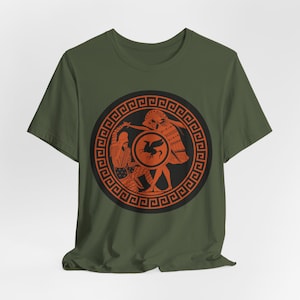 May include: An olive green t-shirt with a circular design. The design features orange Greek figures on a black background, including a warrior, a bird on a shield, and a decorative border. The t-shirt is made of a soft material.
