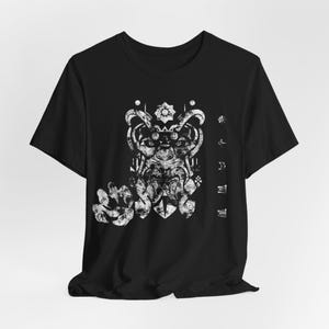 May include: A black short-sleeved t-shirt featuring a distressed white graphic print on the chest. The design depicts a stylised, bearded face with intricate patterns, a star-like emblem above, and a series of small geometric symbols to the right.
