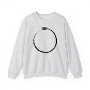May include: A light gray heathered crewneck sweatshirt featuring a black ink-style graphic of an ouroboros, a serpent eating its own tail, centered on the chest. The garment has long sleeves, a ribbed neckline, and ribbed cuffs.