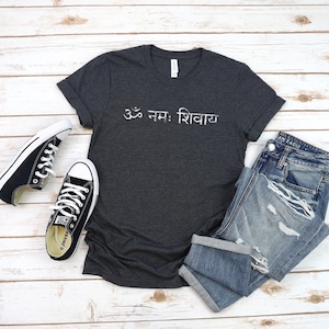 May include: A dark charcoal grey short-sleeve t-shirt featuring the white Sanskrit text Om Namah Shivaya printed across the chest, paired with a pair of black and white low-top canvas trainers and blue denim jeans with distressed detailing.