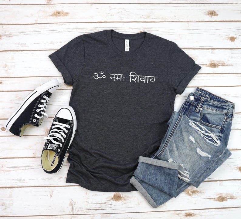 May include: A dark charcoal grey short-sleeve t-shirt featuring the white Sanskrit text Om Namah Shivaya printed across the chest, paired with a pair of black and white low-top canvas trainers and blue denim jeans with distressed detailing.