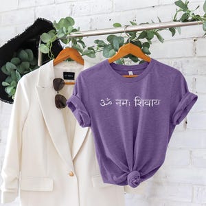 May include: A purple short-sleeved t-shirt featuring the white Sanskrit text Om Namah Shivaya printed across the chest. The shirt has a crew neckline and is displayed on a wooden hanger alongside a white blazer and a black hat.