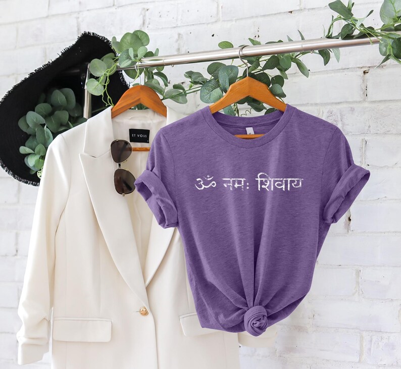 May include: A purple short-sleeved t-shirt featuring the white Sanskrit text Om Namah Shivaya printed across the chest. The shirt has a crew neckline and is displayed on a wooden hanger alongside a white blazer and a black hat.