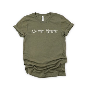 May include: An olive green short-sleeved t-shirt featuring the white text Om Namah Shivaya printed across the chest. The shirt has a crew neckline and rolled sleeves, with a slightly distressed, textured appearance to the white lettering.