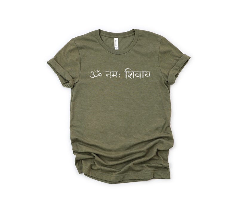 May include: An olive green short-sleeved t-shirt featuring the white text Om Namah Shivaya printed across the chest. The shirt has a crew neckline and rolled sleeves, with a slightly distressed, textured appearance to the white lettering.