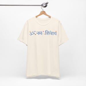 May include: A cream-coloured short-sleeve t-shirt featuring the blue Sanskrit text Om Namah Shivaya printed across the chest. The design includes small, colourful dots interspersed between the characters, all displayed on a wooden hanger.