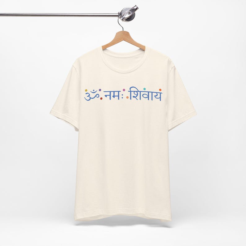 May include: A cream-coloured short-sleeve t-shirt featuring the blue Sanskrit text Om Namah Shivaya printed across the chest. The design includes small, colourful dots interspersed between the characters, all displayed on a wooden hanger.