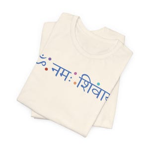 May include: A cream-coloured t-shirt featuring the blue Sanskrit text "ॐ नमः शिवाय" printed across the chest. The design includes small, colourful dots interspersed between the characters, creating a simple and clean visual presentation on the fabric.