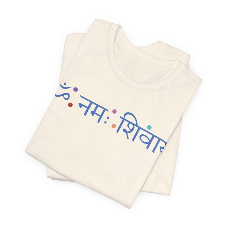 May include: A cream-coloured t-shirt featuring the blue Sanskrit text "ॐ नमः शिवाय" printed across the chest. The design includes small, colourful dots interspersed between the characters, creating a simple and clean visual presentation on the fabric.