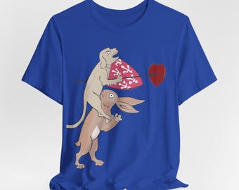 Puede incluir: Una camiseta azul real de manga corta con una ilustración gráfica de un perro marrón claro montado sobre un conejo marrón. El perro sostiene un escudo rojo con marcas blancas, y una flecha negra atraviesa tanto el escudo como un corazón rojo.