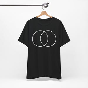 May include: A black short-sleeved t-shirt featuring a white graphic of two overlapping circles centered on the chest. The garment has a crew neckline and is displayed hanging on a wooden hanger against a plain, neutral background.