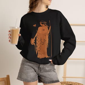 May include: A black long-sleeve sweatshirt featuring a terracotta-colored graphic print of a classical figure in ancient Greek style, holding a spear and a helmet, with a small bird-like shape above the figure's head, worn with gray denim shorts.