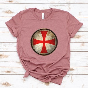 May include: A mauve t-shirt with a distressed, circular graphic of a red cross on a white shield.
