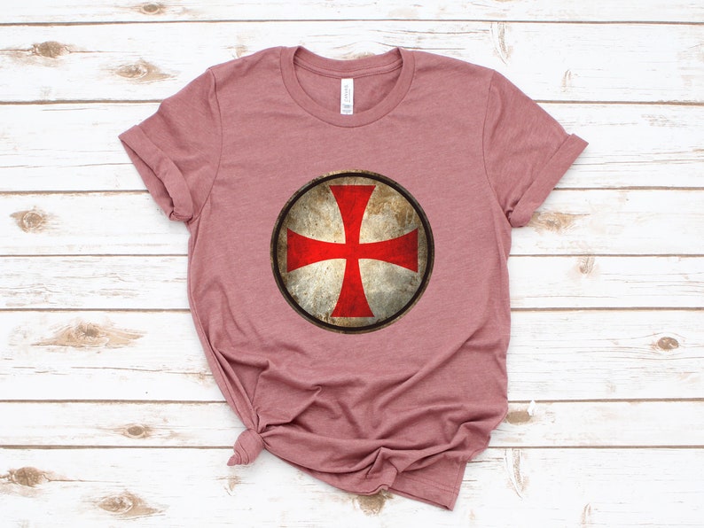 May include: A mauve t-shirt with a distressed, circular graphic of a red cross on a white shield.