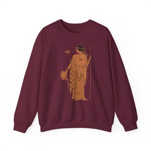 May include: Burgundy sweatshirt with an orange illustration of a classical Greek figure holding a spear and helmet. The design is inspired by ancient pottery art, with a butterfly detail.
