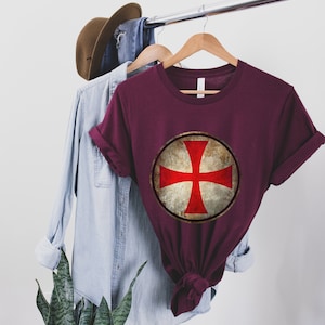 May include: A maroon t-shirt with a vintage-style red cross on a white circle. The cross is a symbol of the Knights Templar.