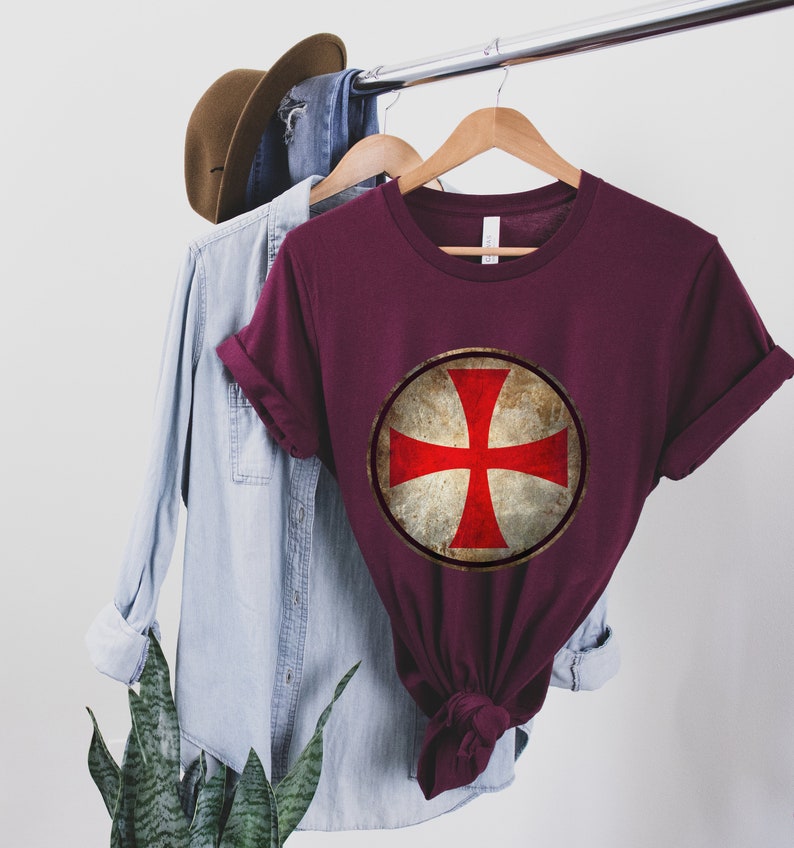 May include: A maroon t-shirt with a vintage-style red cross on a white circle. The cross is a symbol of the Knights Templar.