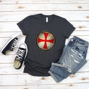 May include: A dark gray t-shirt with a red cross on a white shield. The shield is surrounded by a brown circle with a distressed look. The t-shirt is laid flat on a wooden surface with a pair of black canvas shoes and a pair of blue jeans.
