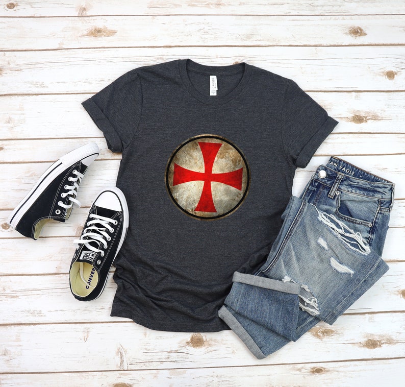 May include: A dark gray t-shirt with a red cross on a white shield. The shield is surrounded by a brown circle with a distressed look. The t-shirt is laid flat on a wooden surface with a pair of black canvas shoes and a pair of blue jeans.