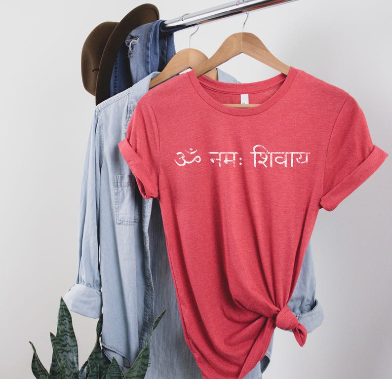 May include: A heathered red short-sleeved t-shirt featuring the white Sanskrit text Om Namah Shivaya printed across the chest. The shirt has a crew neckline and is displayed on a wooden hanger, with a light blue denim shirt and brown hat visible.