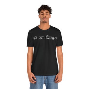 May include: A black short-sleeved t-shirt featuring the white Sanskrit text Om Namah Shivaya printed across the chest. The graphic has a distressed, textured appearance. The shirt is paired with blue denim jeans, completing the casual look.