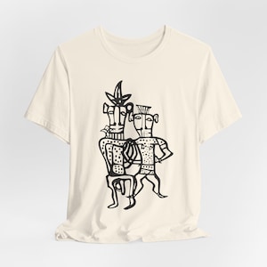 May include: A cream-colored short-sleeve t-shirt featuring a black line-art graphic on the front. The illustration depicts two stylized figures standing side-by-side, characterized by abstract shapes, dotted patterns, and distinct headgear designs.