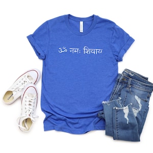 May include: A royal blue short-sleeve t-shirt featuring the white text Om Namah Shivaya in Devanagari script, paired with a pair of white canvas trainers and blue denim jeans with distressed detailing on the legs and frayed hems.