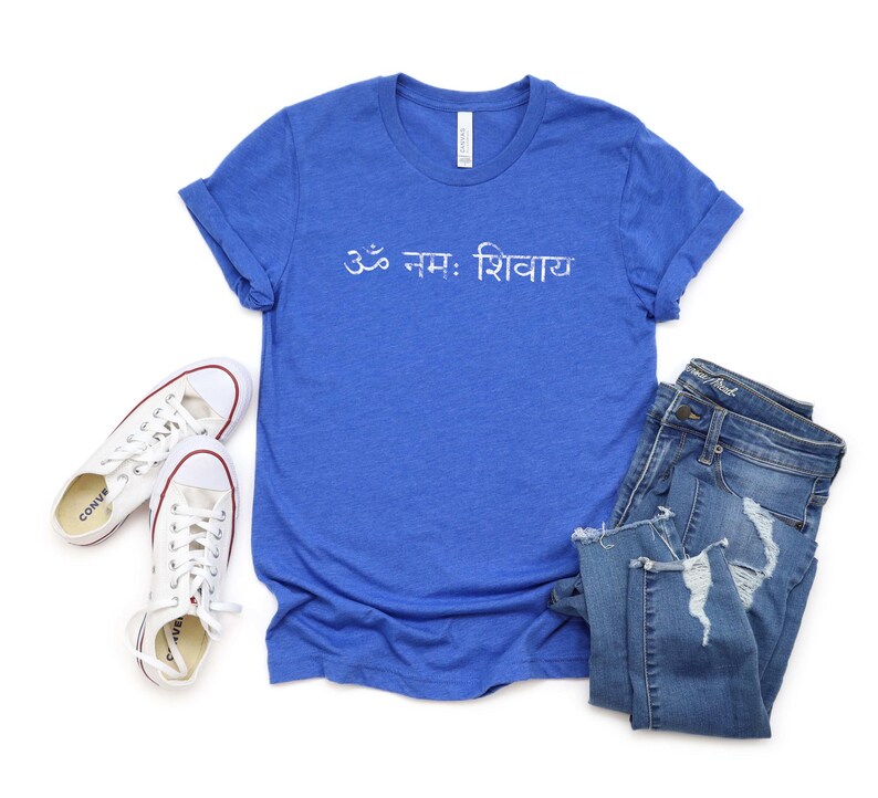 May include: A royal blue short-sleeve t-shirt featuring the white text Om Namah Shivaya in Devanagari script, paired with a pair of white canvas trainers and blue denim jeans with distressed detailing on the legs and frayed hems.